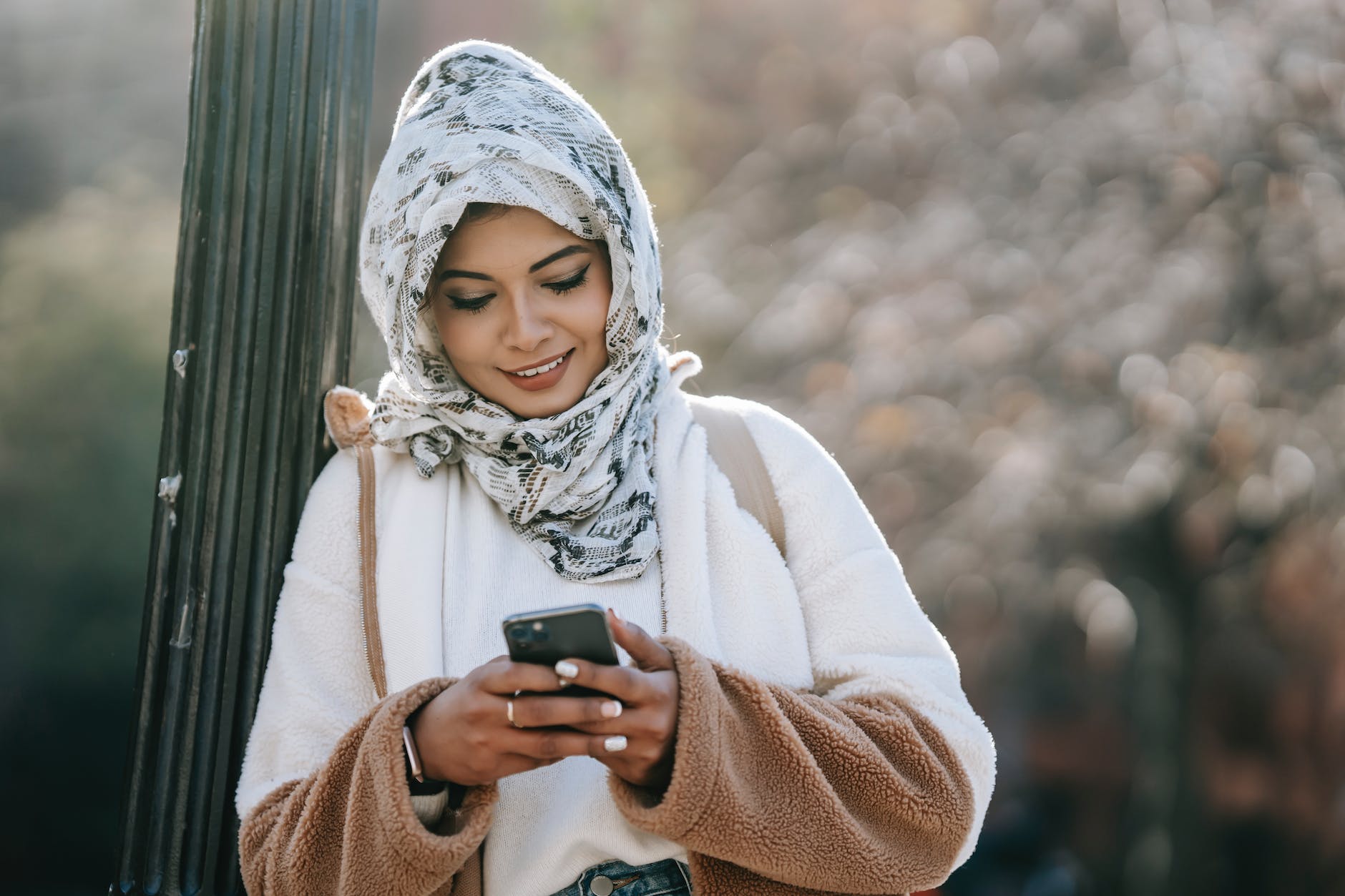cheerful young ethnic female in headscarf and coat browsing smartphone